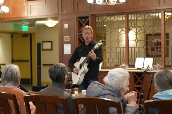 Harmony & Healing musician sings and plays guitar for seniors seated in a care facility common room